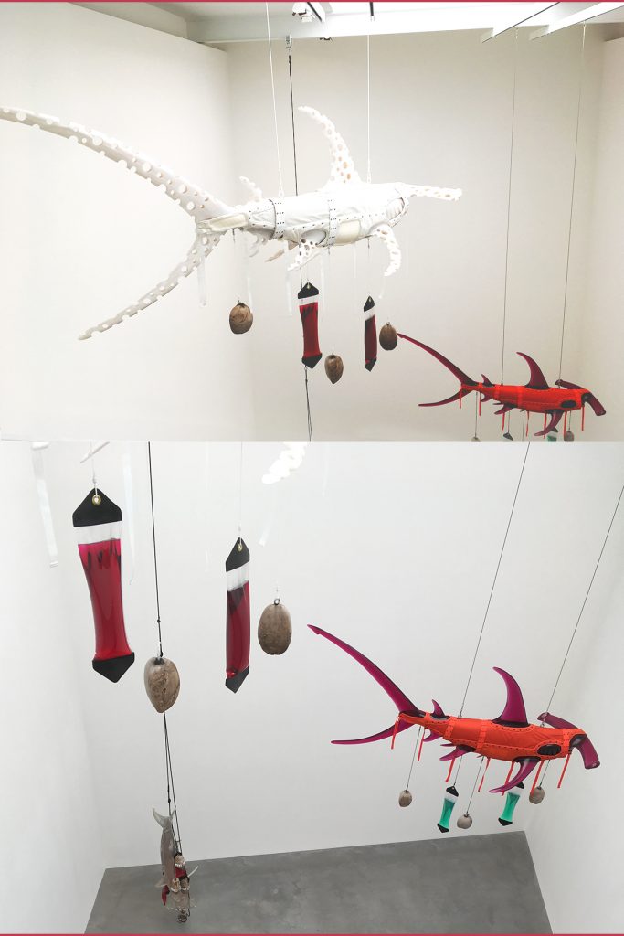 Life size polyurethane resin hammerhead sharks hanging in the gallery, wrapped in nylon webbing, with coconuts and intravenous drip bags hanging from them